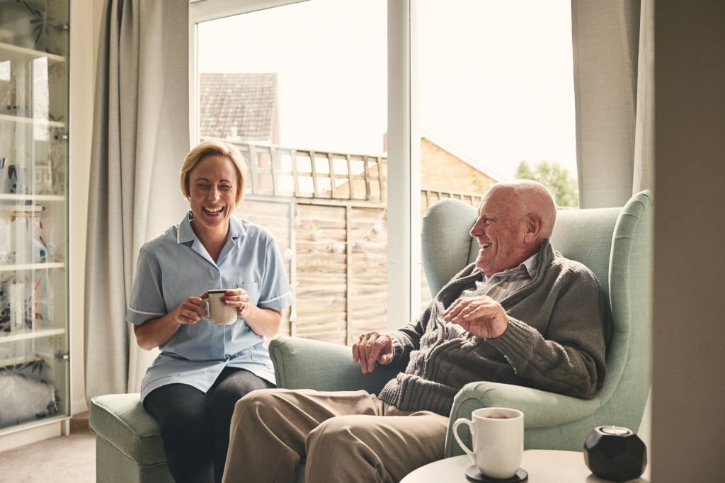 Indoor shot of smiling senior man and female carer enjoying coffee in living room
